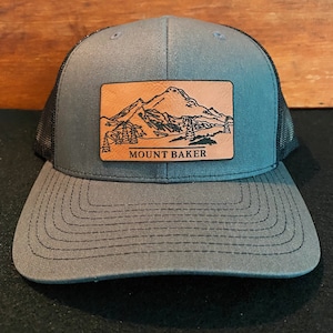 May include: A gray trucker hat with a black mesh back. The hat features a brown leather patch with a black line drawing of Mount Baker and the text "MOUNT BAKER".