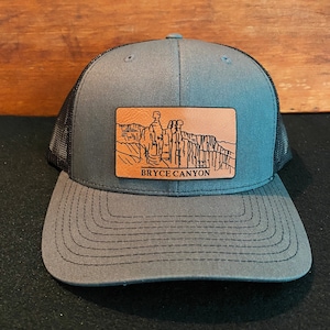 May include: A gray and black trucker hat with a brown leather patch featuring a black and white illustration of Bryce Canyon National Park. The patch has the text "Bryce Canyon" printed on it.