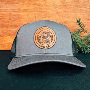 May include: A gray and black trucker hat with a brown leather patch featuring a silhouette of a rock formation and the word "MOAB".