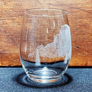May include: A clear glass wine glass with an etched design of a mountain range and trees. The glass is sitting on a dark blue surface.