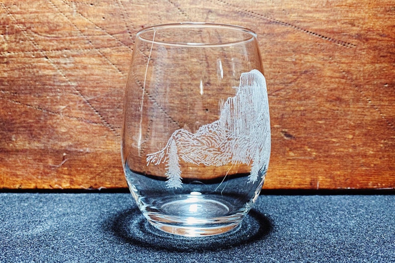 May include: A clear glass wine glass with an etched design of a mountain range and trees. The glass is sitting on a dark blue surface.
