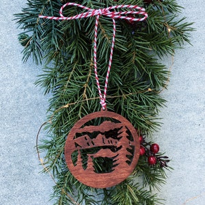 Puede incluir: Un adorno navide&ntilde;o de madera con un dise&ntilde;o recortado de una cordillera y pinos. El adorno es marr&oacute;n y est&aacute; colgado de una cuerda de hilo rojo y blanco.