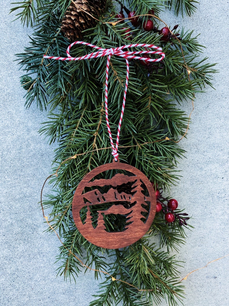Puede incluir: Un adorno navide&ntilde;o de madera con un dise&ntilde;o recortado de una cordillera y pinos. El adorno es marr&oacute;n y est&aacute; colgado de una cuerda de hilo rojo y blanco.
