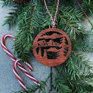 Puede incluir: Un adorno navide&ntilde;o de madera con la silueta de una cordillera de monta&ntilde;as con &aacute;rboles. El adorno es de color marr&oacute;n y tiene una cuerda roja y blanca atada a &eacute;l. El adorno est&aacute; rodeado de ramas de pino verdes.