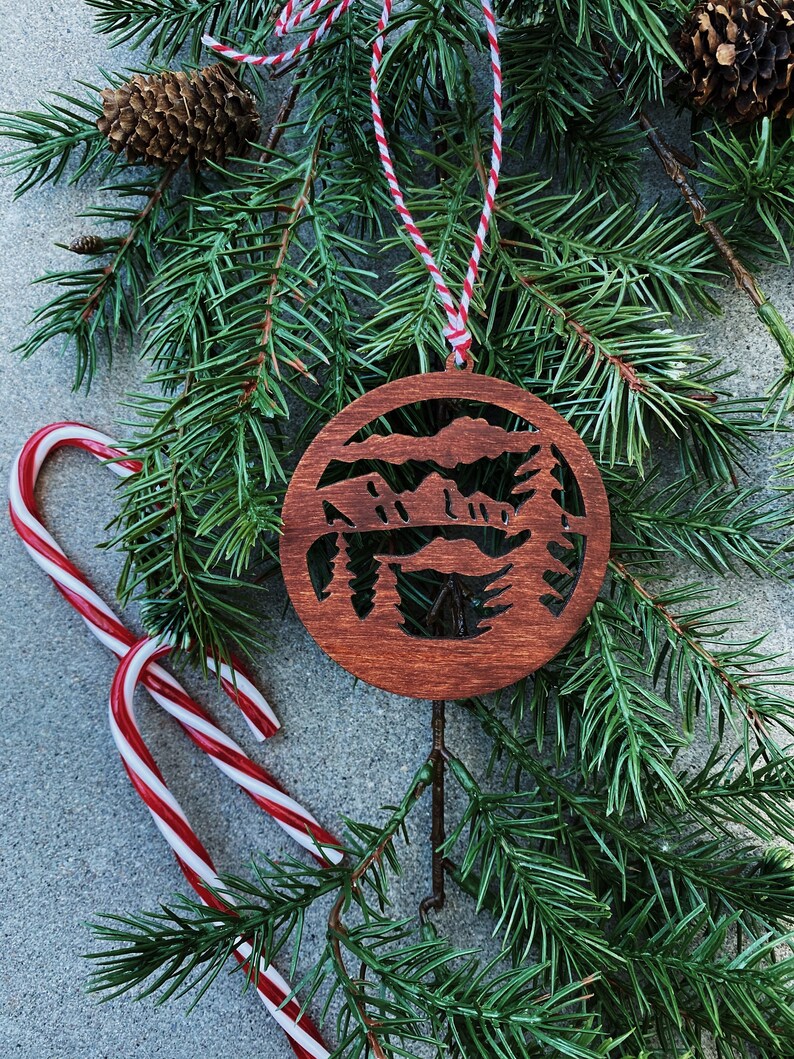 Puede incluir: Un adorno navide&ntilde;o de madera con la silueta de una cordillera de monta&ntilde;as con &aacute;rboles. El adorno es de color marr&oacute;n y tiene una cuerda roja y blanca atada a &eacute;l. El adorno est&aacute; rodeado de ramas de pino verdes.