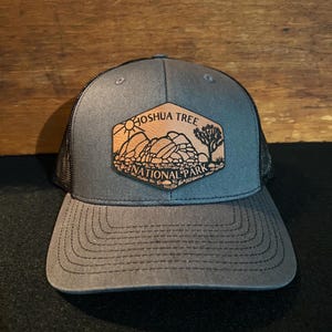 May include: A gray and black trucker hat with a brown leather patch that says "Joshua Tree National Park" with a sun and a Joshua Tree silhouette.