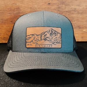 May include: Gray trucker hat with a black mesh back and a curved brim. A rectangular leather patch on the front features a mountain range illustration with the text "SUN VALLEY" in black.