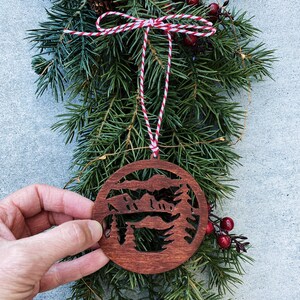 Puede incluir: Un adorno navide&ntilde;o de madera con la silueta de una cordillera de monta&ntilde;as con &aacute;rboles en frente. El adorno es de color marr&oacute;n y tiene un cordel rojo y blanco atado.