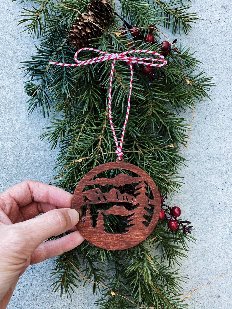 Puede incluir: Un adorno navide&ntilde;o de madera con la silueta de una cordillera de monta&ntilde;as con &aacute;rboles en frente. El adorno es de color marr&oacute;n y tiene un cordel rojo y blanco atado.