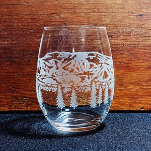 May include: A clear glass wine glass with an etched design of a mountain range and trees. The glass is sitting on a dark surface.