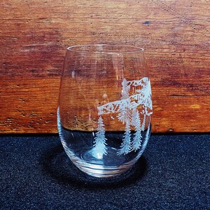 Puede incluir: Una copa de vino de vidrio transparente con un dise&ntilde;o grabado de una escena de bosque. El dise&ntilde;o presenta pinos blancos y es visible a trav&eacute;s del vidrio.