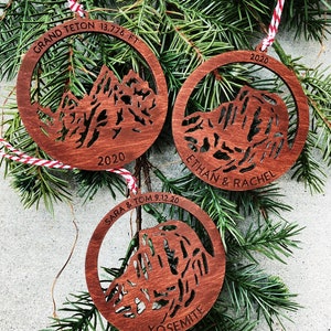 Puede incluir: Tres adornos navide&ntilde;os de madera con siluetas de monta&ntilde;as, personalizados con nombres, fechas y nombres de monta&ntilde;as. Los adornos son marrones con textura de veta de madera. El texto en los adornos dice "GRAND TETON 13,776 FT 2020", "ETHAN & RACHEL 2020" y "SARA & TOM 9.12.20 YOSEMITE".
