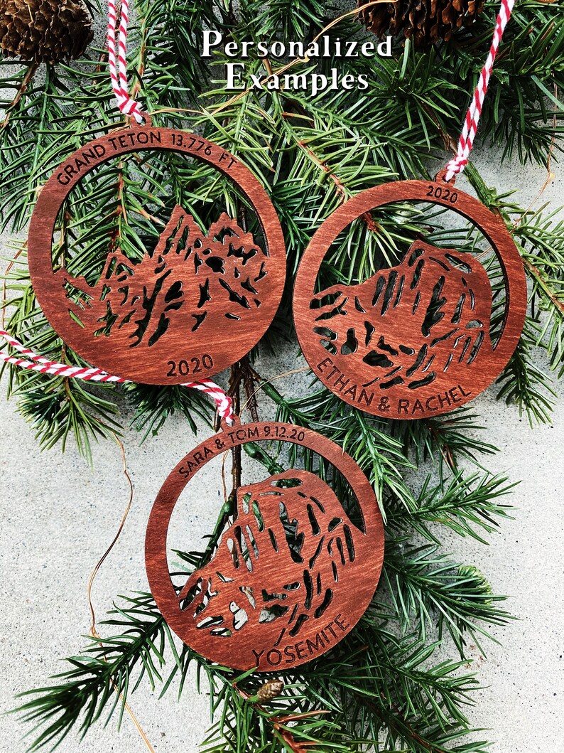 Puede incluir: Tres adornos navide&ntilde;os de madera con siluetas de monta&ntilde;as, personalizados con nombres, fechas y nombres de monta&ntilde;as. Los adornos son marrones con textura de veta de madera. El texto en los adornos dice "GRAND TETON 13,776 FT 2020", "ETHAN & RACHEL 2020" y "SARA & TOM 9.12.20 YOSEMITE".