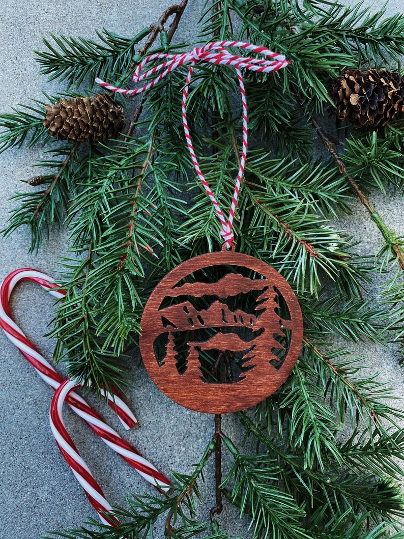 Puede incluir: Un adorno navide&ntilde;o de madera con un dise&ntilde;o de corte l&aacute;ser de una escena de monta&ntilde;a con &aacute;rboles. El adorno es marr&oacute;n y tiene un colgador de cuerda roja y blanca.