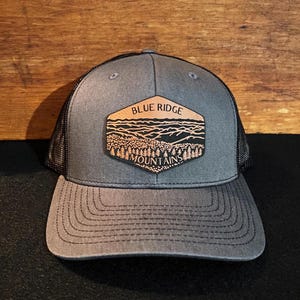 May include: A gray and black trucker hat with a brown leather patch that says "Blue Ridge Mountains" with a mountain range design.