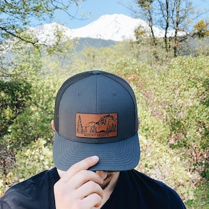 May include: A gray and black trucker hat with a brown leather patch featuring a mountain scene and the text "MOUNT SHASTA".