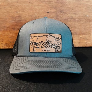 May include: A gray and black trucker hat with a brown leather patch featuring a mountain range and the word "ASPEN".