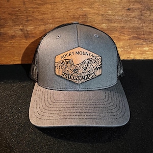 Peut inclure: Un chapeau de camionneur gris et noir avec un patch en cuir brun qui dit "Rocky Mountain National Park" avec une illustration de chaîne de montagnes.