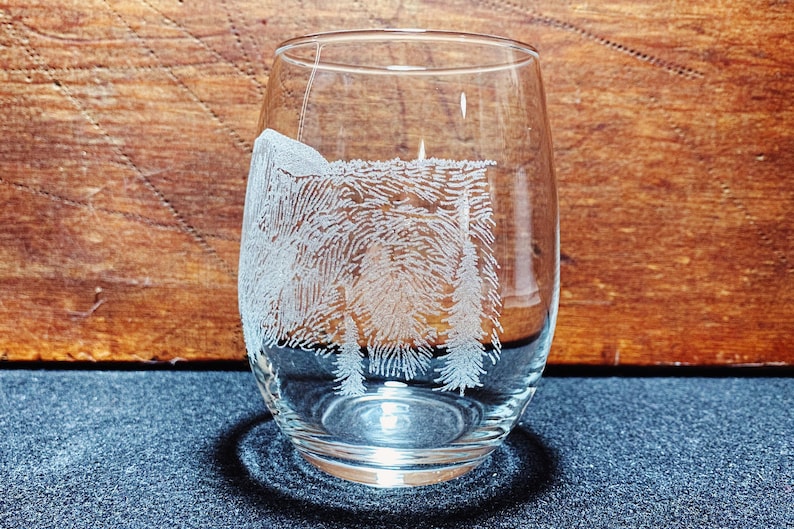 May include: A clear glass wine glass with an etched design of a forest scene. The design features trees and a mountain in the background.