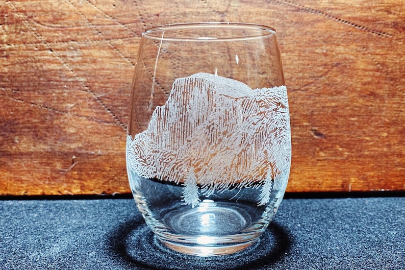 May include: A clear glass wine glass with a white etched design of a mountain range. The glass is sitting on a dark blue surface.