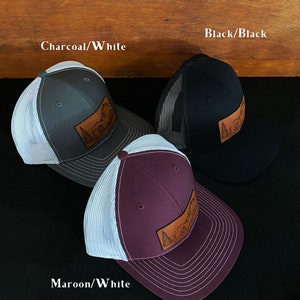May include: Three trucker hats with a leather patch featuring a mountain scene. The hats are in charcoal/white, black/black, and maroon/white.