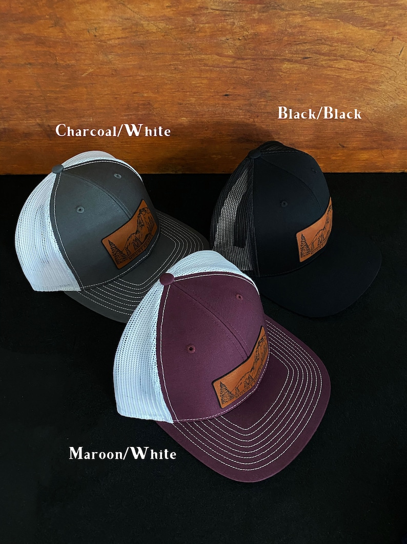 May include: Three trucker hats with a leather patch featuring a mountain scene. The hats are in charcoal/white, black/black, and maroon/white.