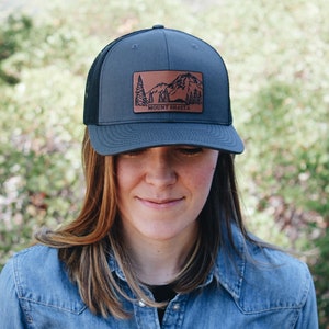 May include: A gray and black trucker hat with a brown leather patch featuring a mountain range and the text "MOUNT SHASTA".