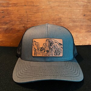 May include: A gray and black trucker hat with a brown leather patch featuring a black and white illustration of Yosemite National Park and the word "Yosemite".