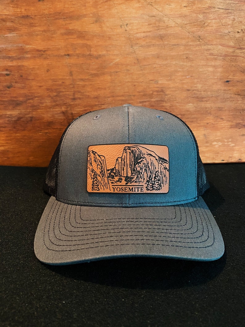 May include: A gray and black trucker hat with a brown leather patch featuring a black and white illustration of Yosemite National Park and the word "Yosemite".