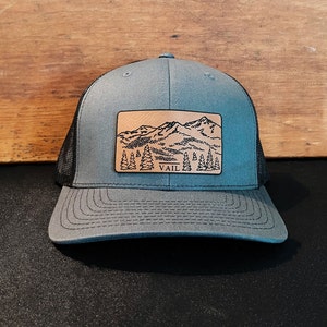 May include: A grey and black trucker hat with a brown leather patch featuring a mountain range and the word "VAIL" in white.