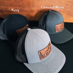 May include: Three trucker hats with black mesh back panels and leather patches featuring a mountain scene. The hats are in navy, charcoal, and heather gray.