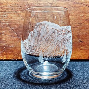 May include: A clear glass wine glass with an etched design of a mountain range. The glass is sitting on a dark surface.