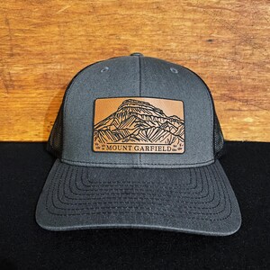 May include: A dark gray trucker hat with a black mesh back. The hat features a brown leather patch with a black outline of Mount Garfield and the text "MOUNT GARFIELD".