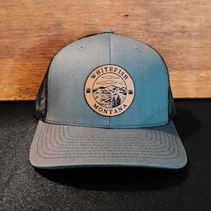 May include: A gray and black mesh trucker hat with a brown leather patch that says "Whitefish Montana" and features a mountain scene with a bear paw print on each side.