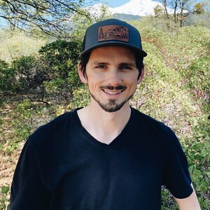 May include: A man wearing a black t-shirt and a black and brown baseball cap with a brown patch that says "Mount Shasta".