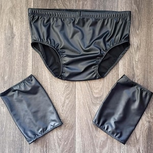 Op de afbeelding: Zwarte ondergoedset met bijpassende armmanchetten. Het ondergoed heeft een elastische tailleband en beenopeningen. De armmanchetten zijn rechthoekig en hebben ook elastische openingen. De set is gemaakt van een glanzend, glad materiaal.