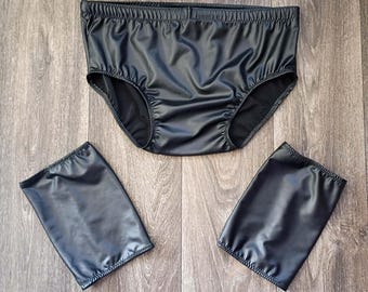 May include: Black underwear set with matching arm cuffs. The underwear has an elastic waistband and leg openings. The arm cuffs are rectangular and also have elastic openings. The set is made of a shiny, smooth material.