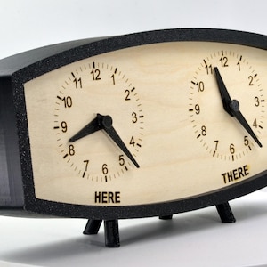 May include: A dual-dial clock with a black frame and wooden face. The clock has two faces, each with black hands and numbers. The word "HERE" is below one clock face, and "THERE" is below the other.