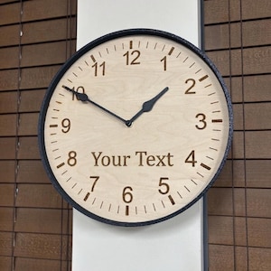 May include: A round wall clock with a black frame and a light wood face. The clock features black numerals from 1 to 12, black hour and minute hands, and the text "Your Text" engraved in the center. The design includes black tick marks.