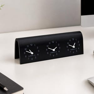 以下が含まれることがあります： マットブラックのアーチ型デスククロックで、3つのアナログ時計の文字盤が水平に並んでいます。各文字盤には白い数字と白い針があり、中央の下にLabel-1、Label-2、Label-3というテキストがそれぞれ印刷されています。