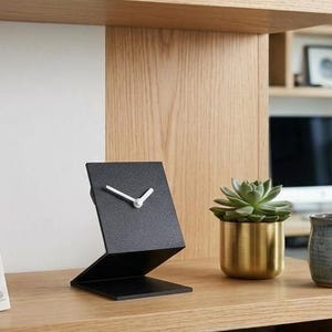 May include: A black, Z-shaped desk clock with a textured finish and white hands. The clock is positioned on a wooden surface next to a green succulent in a gold-toned metal pot and a grey ceramic mug containing two pens.