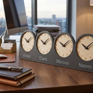 May include: A desk clock featuring four circular clock faces with light wood grain backgrounds and black hands, set into a dark gray textured base. The names Julian, Clara, Marcus, and Naomi are engraved in white text below each respective clock face.