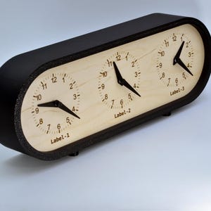May include: A black oval-shaped clock with three analog clock faces. Each clock face has a white background with black numbers and hands. The clock faces are labeled "Label-1", "Label-2", and "Label-3".