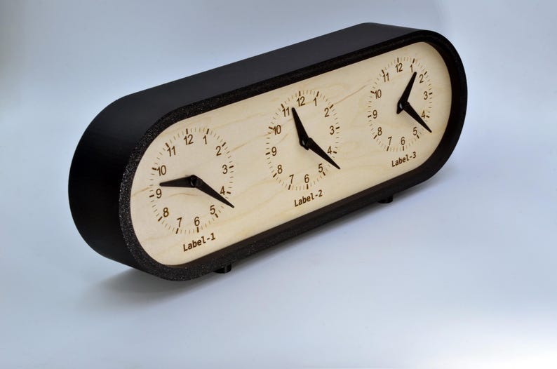 May include: A black oval-shaped clock with three analog clock faces. Each clock face has a white background with black numbers and hands. The clock faces are labeled "Label-1", "Label-2", and "Label-3".