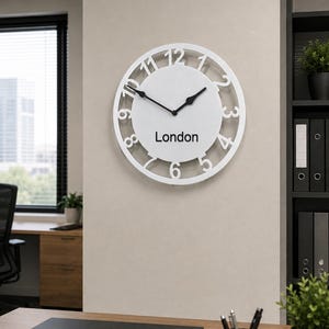 May include: A circular wall clock with a white frame and a white face featuring large, cut-out black numbers from one to twelve. The clock has two black hands and the word London printed in black text in the centre of the clock face.