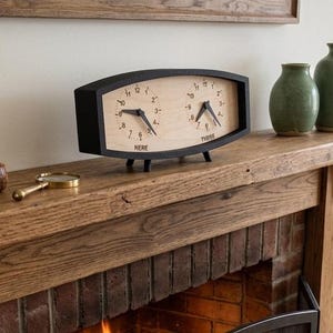 May include: A dual-faced mantle clock with a black frame and a light wood grain face. The left clock face is labeled "HERE" and the right clock face is labeled "THERE". Each face features black numbers and black clock hands, supported by small black feet.