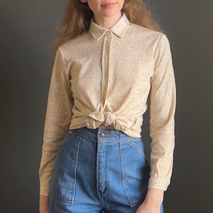 May include: A long-sleeved button-down shirt in a cream color with a fine brown speckled pattern, tied at the waist, paired with high-waisted blue denim jeans featuring large front patch pockets and a central metal button closure.