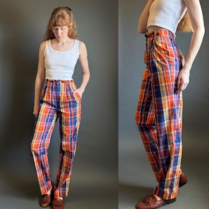 May include: High-waisted trousers featuring a vibrant plaid pattern in shades of orange, blue, yellow, and white. These pants include a button closure at the waistband, front pleats, and side pockets, paired with a simple white ribbed tank top.