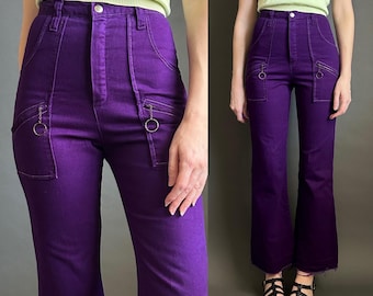 Könnte beinhalten: Hoch taillierte, violette Jeanshose mit ausgestelltem Bein, Knopf- und Reißverschluss sowie zwei Vordertaschen, die mit silberfarbenen Metallringen an kurzen Ketten verziert sind. Die Hose hat einen rohen, ungesäumten Beinabschluss.