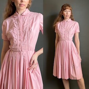 May include: A light pink dress featuring a rounded collar, short sleeves with cuffs, and a button-down bodice with vertical pleats. The skirt is pleated and falls to knee length, creating a structured silhouette with a clean, solid color finish.